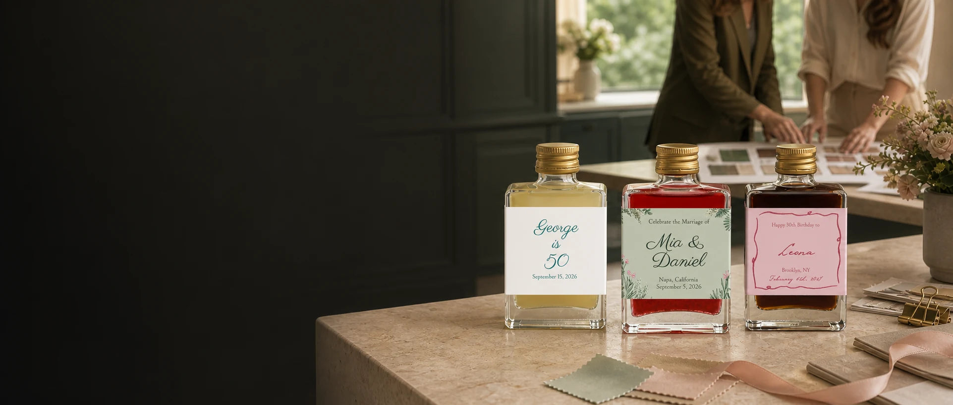 Three personalized Maux cocktail bottles on a design studio counter while two event planners review label proofs and swatches behind them